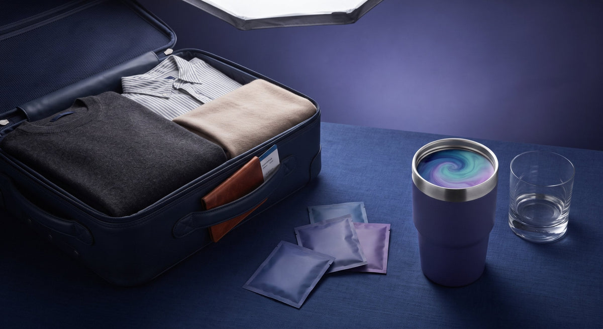 Open suitcase packed for travel with folded clothes, a color-shift travel mug showing a teal and lavender swirl, three unmarked mushroom coffee packets, and a passport on a dark blue table