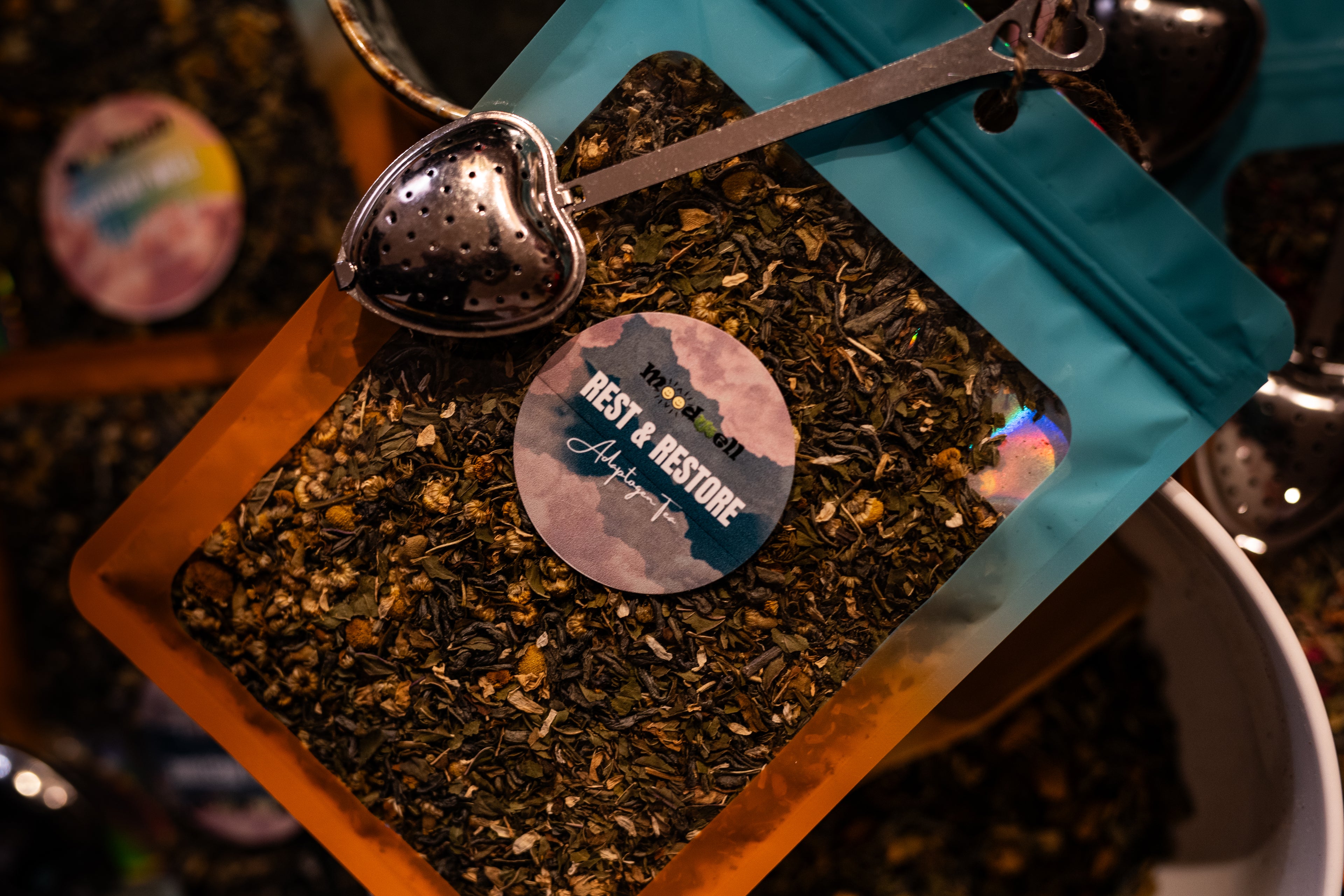 A package of Rest and Restore Adaptogen Tea with a label showing the brand and product name, hanging from a spoon with a background of other tea packages.